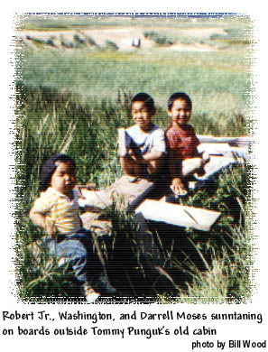 Robert Jr., Washington, and Darrell Moses, suntanning on a board outside tommy Punguks old cabin. The old airport runway and Golovnin Bay are visable in the distance. Photo (c) Bill Wood